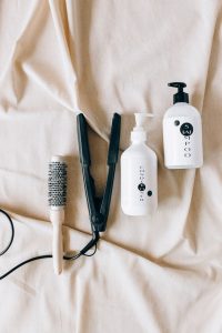 A round hair brush, a flat iron and a labeled bottle of shampoo and conditioner.